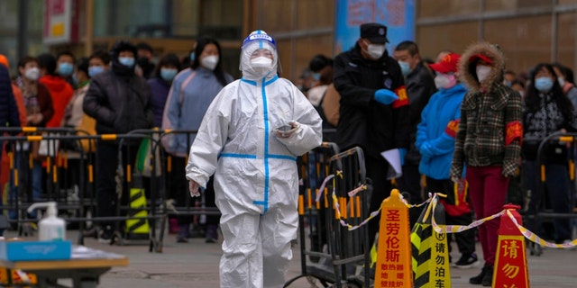 A health worker wearing a protective suit walks by masked residents who wait in line to get their throat swab at a coronavirus testing site after a COVID-19 case was detected in a residential building, Wednesday, April 6, 2022, in Beijing.