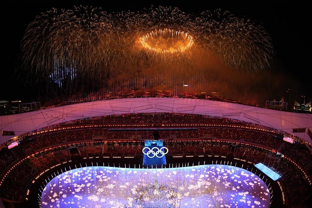 A shower of fireworks, with a golden ring in the center, is seen over a stadium bearing the Olympics' five-ring logo