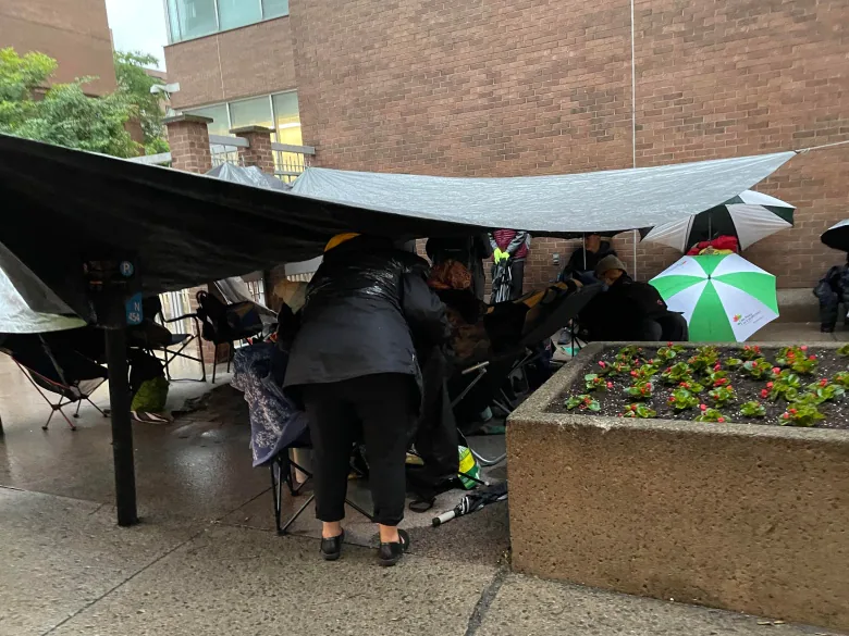 A large tarp is held up by ropes above people waiting in line outside a Montreal passport office. 