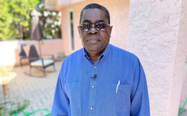 A man standing outside a home, wearing a blue shirt and sunglasses, looks at the camera.