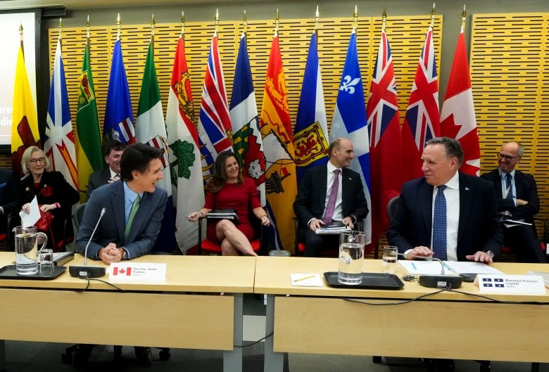 Two smiling men wearing suits sit at a long table with microphones, in front of a row of seated people under a wall with large Canada and provincial flags.