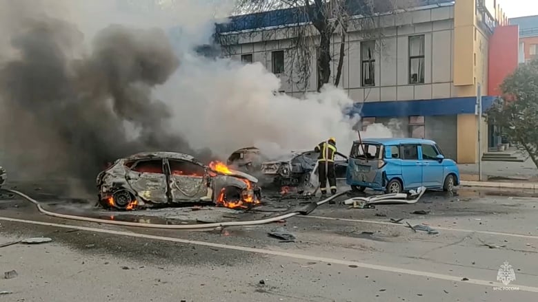 A firefighter works to extinguish burning cars on a street. Smoke billows from the vehicles.