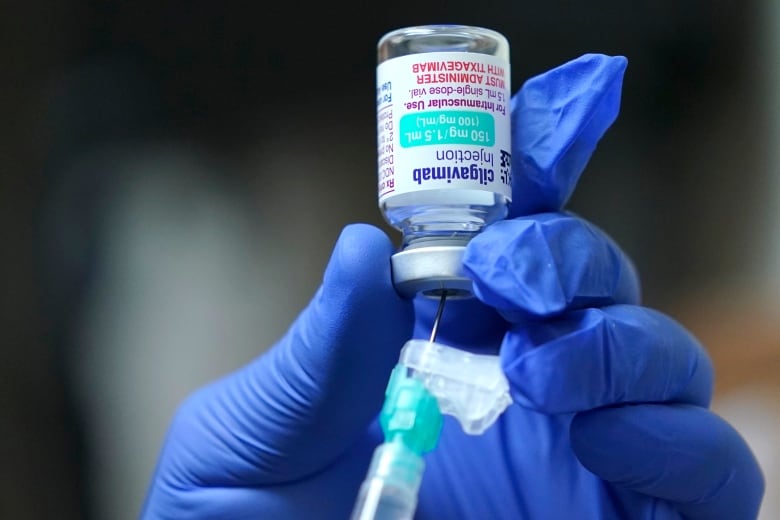 A hand in a blue latex glove holds a small upside-down vial and inserts a syringe into it.