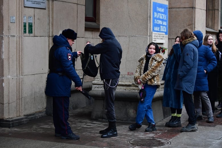 A police officer with a handheld metal detector inspects a person's backpack as a line of people wait in a line.