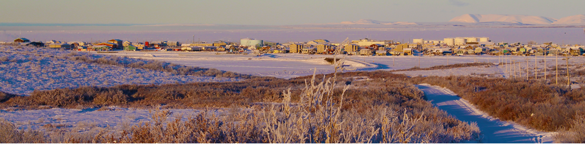 A panoramic view of Alaska's Northwest Arctic Borough. In the foreground is frozen ground covered in snow and ice; in the background are various buildings. Pink and white clouds streak across the sky. 