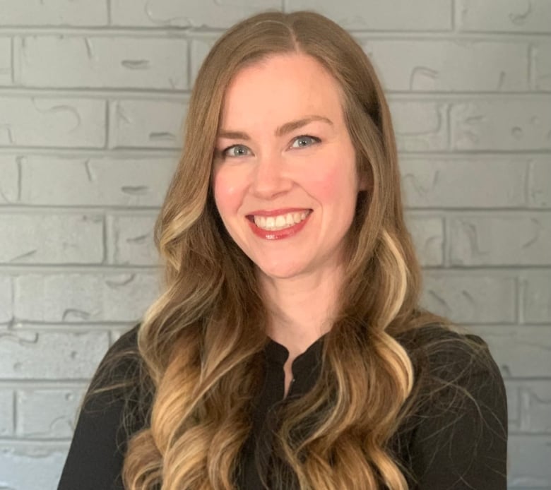 A woman with long light brown hair and blue eyes stands in front of a light coloured brick wall with her arms crossed. She is wearing a long sleeve black shirt.