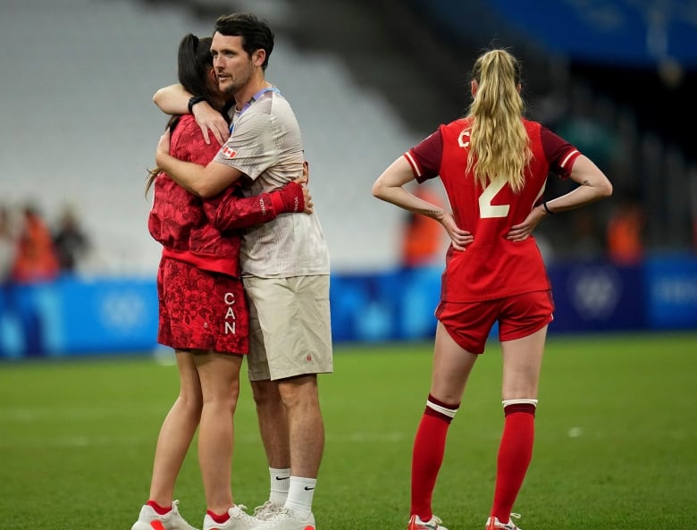 A man wearing Team Canada clothes hugs a female soccer player on a soccer pitch, while another player looks onward.