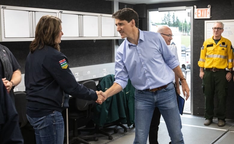 Prime Minister Justin Trudeau meets with Alberta Premier Dannielle Smith. They are shaking hands and saying something to one another. Trudeau is in a blue button up and blue jeans, while Smith is wearing a black jacket and blue jeans. They are in a small room with dark walls and fluorescent lighting.