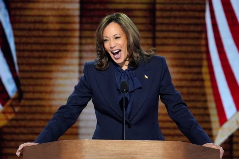 A smiling woman speaks at a podium flanked by American flags.