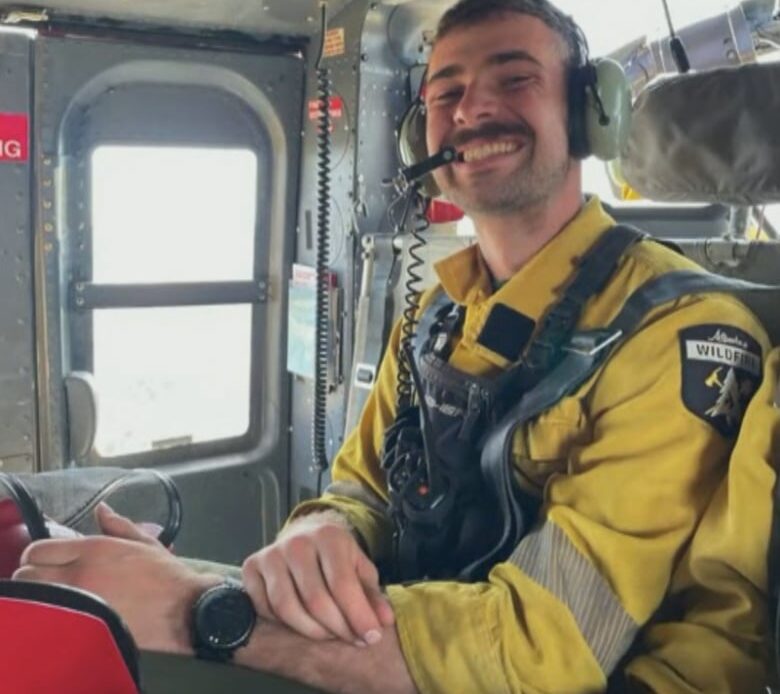 A firefighter with short brown hair and yellow jumpsuit smiles while taking a trip in a helicopter.