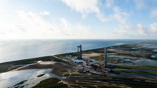 long-distance view of a launch tower holding a silver rocket above the ground, with the ocean in the background and wetlands around the launch pad