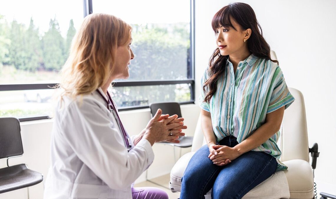 Female patient is shown sat on a white chair at the doctor's office. She has her hands crossed on top of her lap and is looking at a female doctor who appears to be explaining something to her.