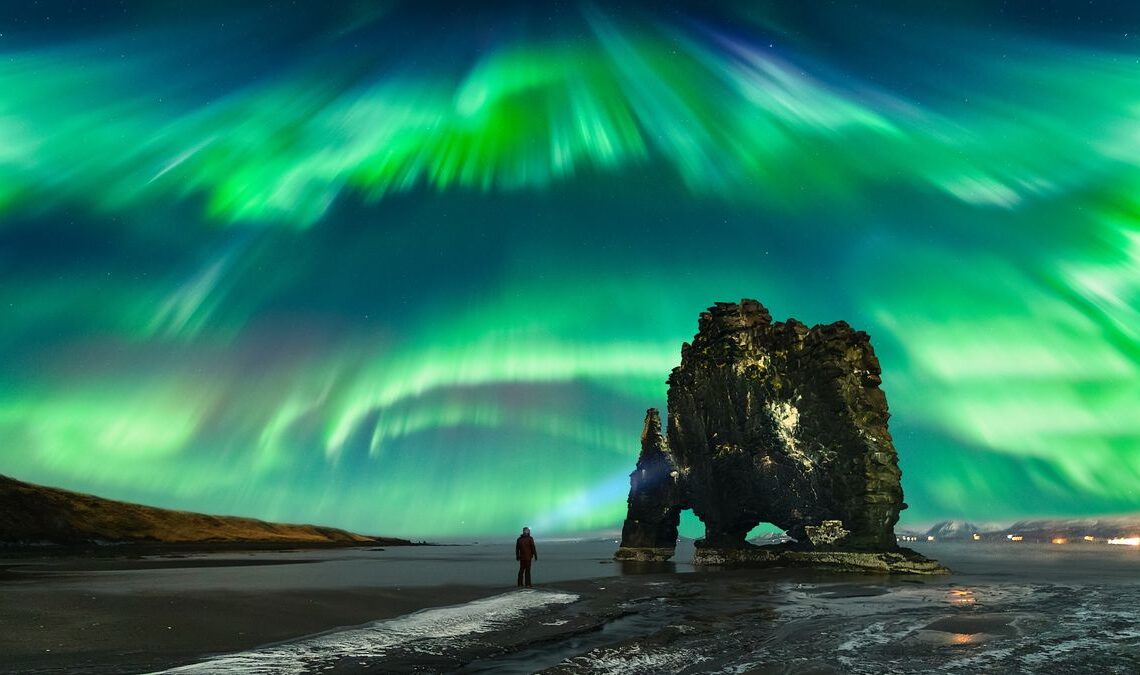 A person stands on a dark beach under a vibrant display of green and blue northern lights, which streak across the sky in a dynamic, swirling pattern. A large, rugged rock formation rises nearby, adding to the dramatic scene.