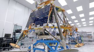 a cube-shaped spacecraft covered in gold foil sits on a dolly in a white cleanroom