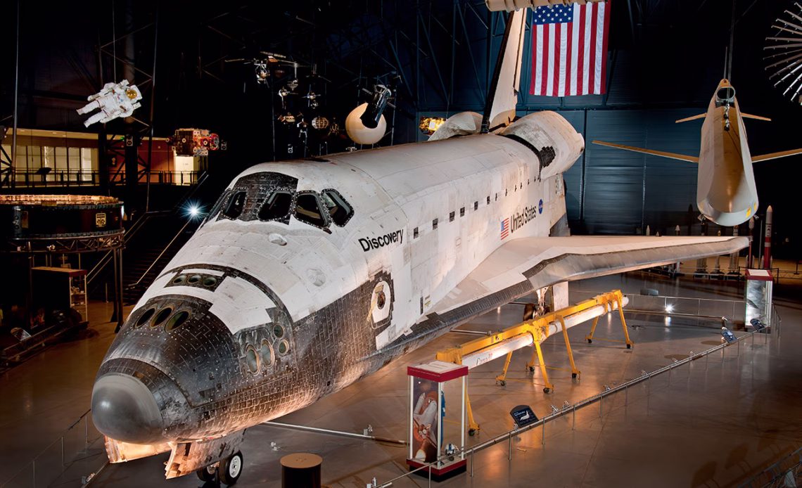 A large white space shuttle sits in a darkened room with the American flag above it.