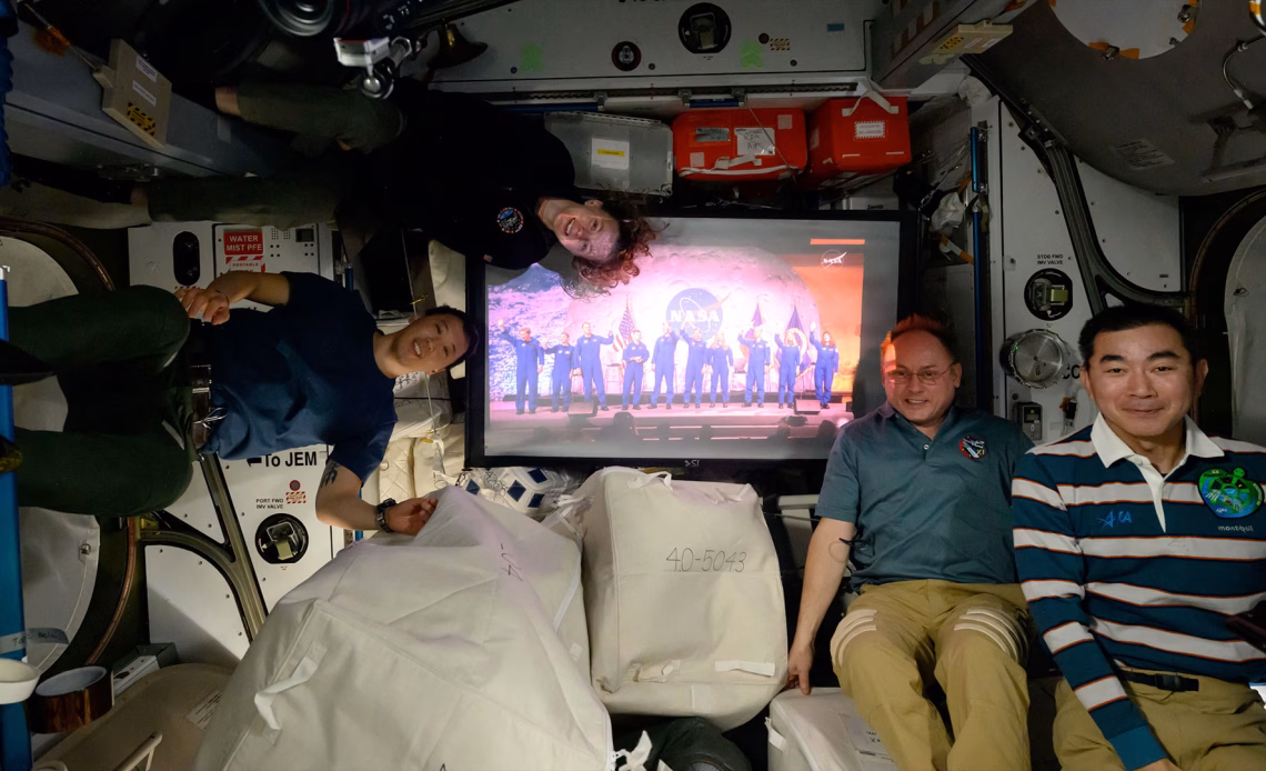 three men and a woman in polo shirts and slacks float on a space station near a screen showing 10 people in blue flights.