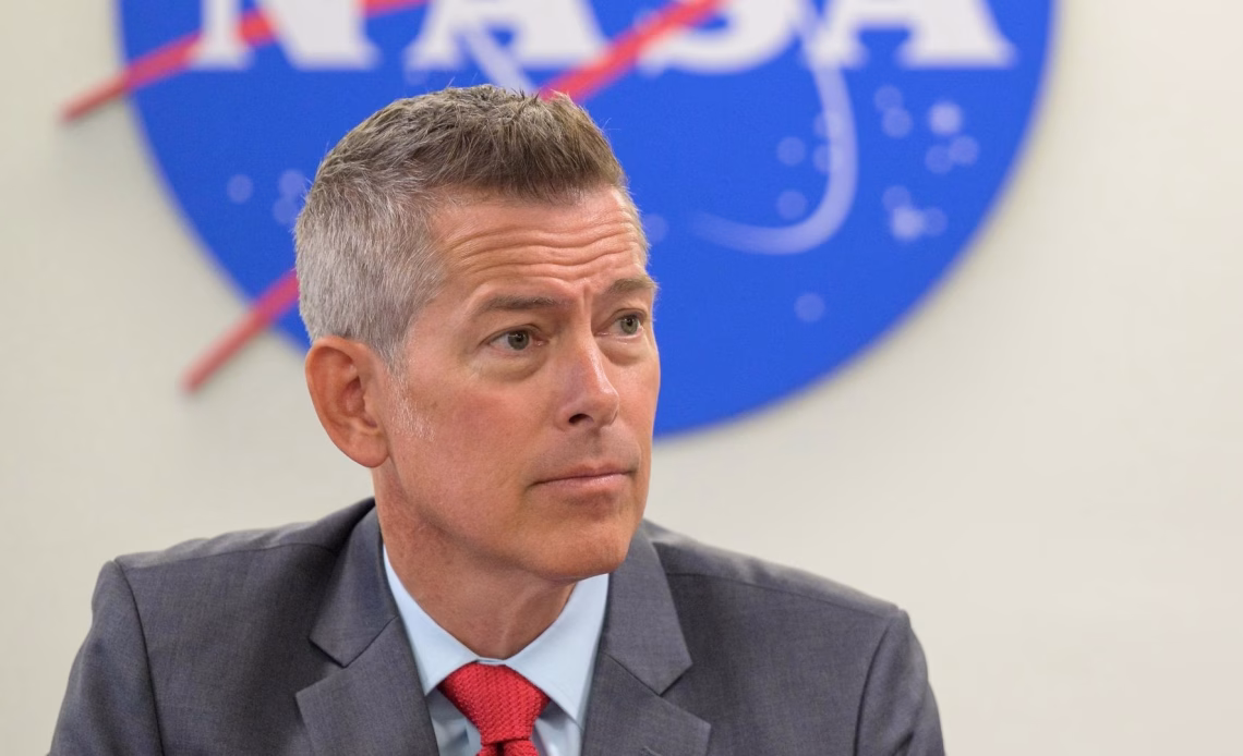 A man in a suit sit looks to the right in front of the bottom half of a nasa meatball logo.