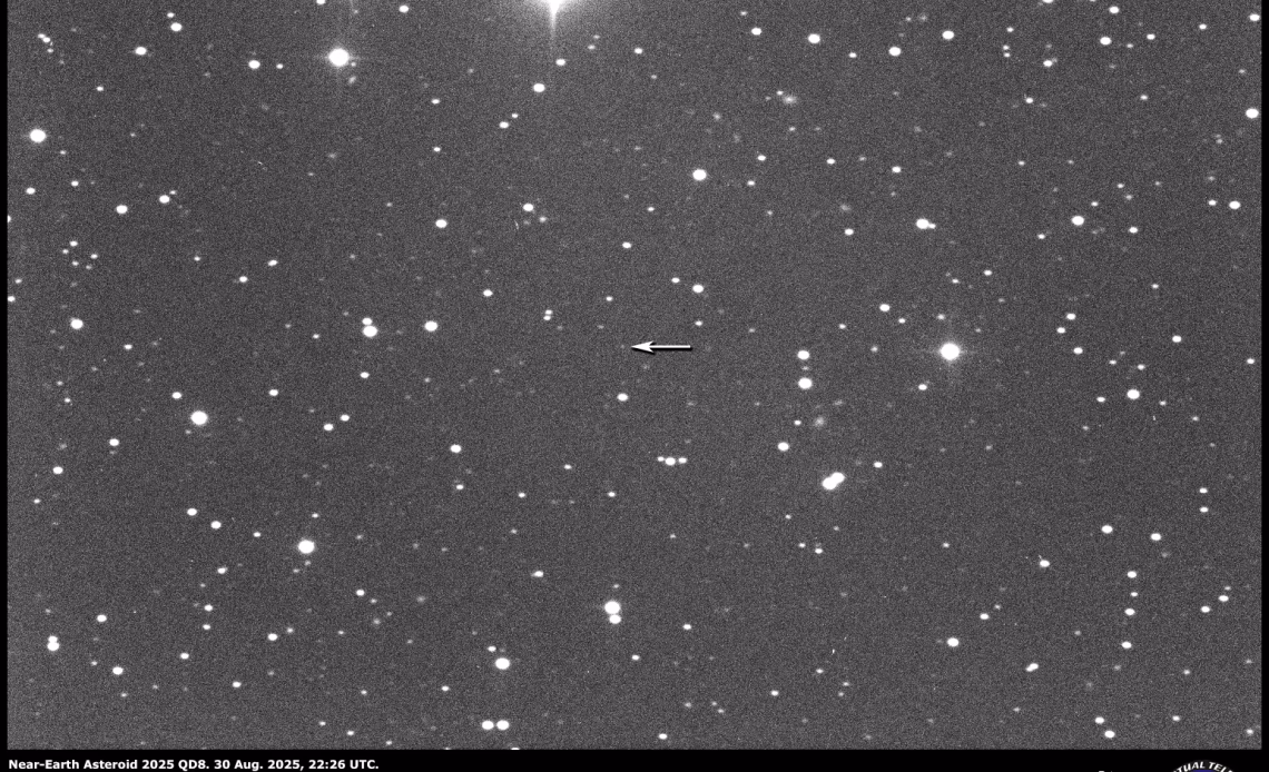White points of light representing stars are visible against a dark background. A white arrow points to the position of a point of light representing a distant asteroid.